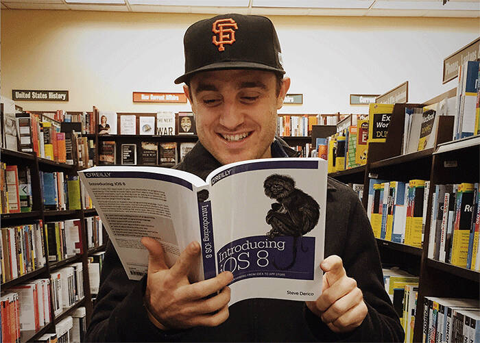 A man wearing a black San Francisco Giants baseball cap smiling while reading the book 'Introducing iOS 8' by Steve Derico in a bookstore.
